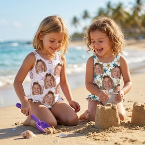 May include: Two young girls on a beach wearing matching white swimsuits. The swimsuits are printed with a repeating pattern of a smiling face, seashells, and starfish. One girl is using a purple shovel to play in the sand.