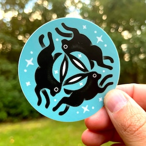 May include: A round sticker with a blue background and three black rabbits in a circle. The rabbits are facing inwards and their ears are overlapping. There are white stars scattered around the rabbits.