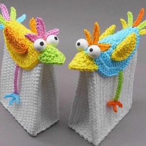 Könnte beinhalten: Zwei weiße gehäkelte Taschen mit bunten vogelartigen Aufsätzen. Die Vögel haben große Wackelaugen und leuchtende gelbe, orangefarbene, blaue, grüne und rosa Akzente. Einer hat eine rosa und blaue Schnur, der andere eine grüne und orangefarbene.