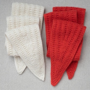 Könnte beinhalten: Nahaufnahme von vier gestrickten Schals. Zwei sind elfenbeinfarben und zwei sind rot. Jeder Schal hat eine dreieckige Form und eine gerippte Textur. Die Schals sind auf einer hellgrauen, strukturierten Oberfläche angeordnet.