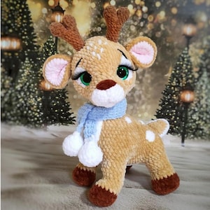Puede incluir: Un juguete de reno de peluche hecho a mano con cuernos marrones, una bufanda azul claro y manchas blancas. El reno tiene grandes ojos verdes, largas pestañas y orejas forradas de rosa. El juguete está sobre una superficie con un fondo borroso de escena invernal.