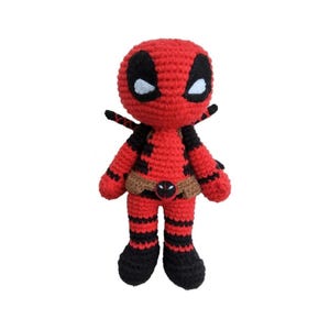 Könnte beinhalten: Handgefertigte Deadpool-Puppe aus Häkelarbeit. Die Puppe ist hauptsächlich rot mit schwarzen Akzenten, einschließlich der Maske, Streifen und Stiefel. Sie hat einen braunen Gürtel mit Deadpool-Logo und kleine schwarze Flügel.