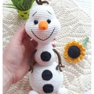 May include: A crocheted snowman toy with a carrot nose, black eyes, and three black buttons. It has brown twig arms and hair. A small crocheted sunflower is next to the snowman.