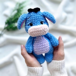 May include: A handmade blue crochet donkey plush toy with a light purple belly and a black bow. The donkey has a light pink muzzle and inner ears. The plush toy is held in a person's hands.