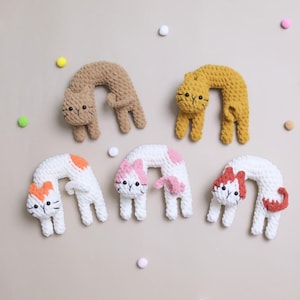 Może przedstawiać: Sześć ręcznie robionych zabawek dla kotów zrobionych na szydełku w różnych kolorach. Koty są wygięte, ze szczegółowymi twarzami i małymi łapkami. Kolory obejmują beżowy, żółty, biały z pomarańczowymi plamkami, biały z różowymi plamkami i biały z czerwonymi akcentami. Rozrzucone są dekoracyjne pompony.