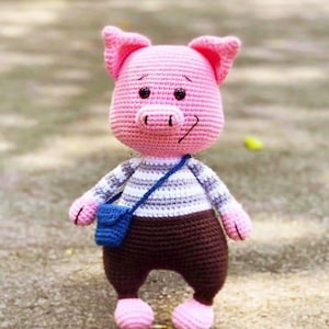 Puede incluir: Una muñeca de cerdo de ganchillo hecha a mano con cabeza, orejas y pies rosas. Lleva una camisa a rayas, pantalones marrones y un bolso cruzado azul. El cerdo tiene ojos negros y una sonrisa cosida.