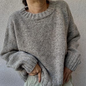 Pode incluir: Um suéter cinza claro com gola redonda e gola e punhos canelados. O suéter tem um ajuste solto e confortável e é feito de um tecido de malha texturizado. As mangas são compridas e o estilo geral é casual e descontraído.