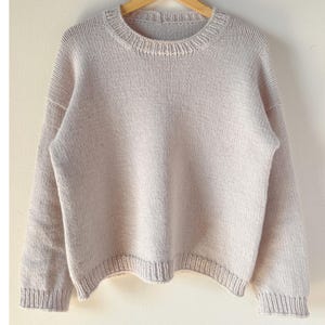 Könnte beinhalten: Hellbeige Pullover mit Rundhalsausschnitt und langen Ärmeln. Der Strickpullover hat Rippendetails an den Bündchen, am Saum und am Ausschnitt. Das Kleidungsstück hängt an einem Holzkleiderbügel.