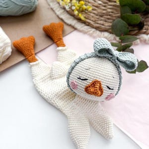 Könnte beinhalten: Ein handgefertigtes, weißes Häkelgans-Spielzeug mit orangefarbenen Füßen und Schnabel sowie einer hellblauen Schleife. Die Gans hat geschlossene Augen und rosa Wangen. Das Spielzeug liegt auf einer weißen und rosa Oberfläche, mit Garn und Blumen im Hintergrund.