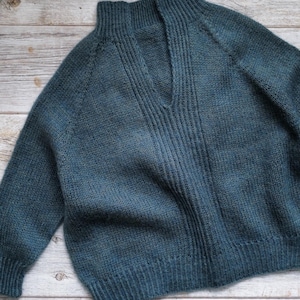 Puede incluir: Un suéter tejido a mano de color verde azulado con un diseño de cuello en V. El suéter presenta una textura acanalada en la parte delantera y un ajuste relajado. Las mangas son largas y el escote está ligeramente elevado. El hilo tiene una apariencia sutil y jaspeada.