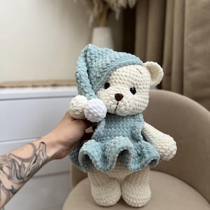 Puede incluir: Un oso de peluche hecho a mano, crema y azul claro. El oso lleva un gorro de dormir azul claro con dos pompones blancos y un vestido a juego. El oso tiene una nariz marrón y ojos negros, y está hecho de hilo suave y texturizado.