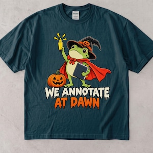 May include: A dark teal short-sleeve t-shirt featuring a graphic of a green frog wearing a witch hat and red cape, holding a yellow highlighter and a book next to a jack-o'-lantern. The text "WE ANNOTATE AT DAWN" is printed in white and orange.