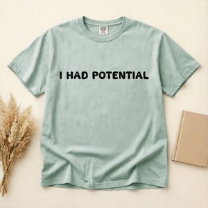 Puede incluir: Una camiseta de manga corta en color verde salvia claro con cuello redondo. En el centro del pecho presenta el texto negro "I HAD POTENTIAL" en una fuente redondeada y negrita. La prenda incluye una etiqueta interior "COMFORT COLORS".