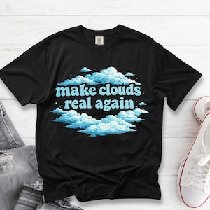 Puede incluir: Camiseta negra con la frase "make clouds real again" en azul claro, rodeada de gráficos de nubes. La camiseta se combina con vaqueros y zapatillas blancas, lo que sugiere un uso informal.