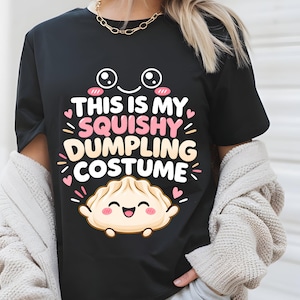 Könnte beinhalten: Ein schwarzes T-Shirt mit kurzen Ärmeln und einer Grafik eines lächelnden Teigtäschchens mit rosa Wangen und kleinen Armen. Der Text THIS IS MY SQUISHY DUMPLING COSTUME steht in weißen und rosa Buchstaben, umgeben von kleinen rosa Herzen und Strichen.