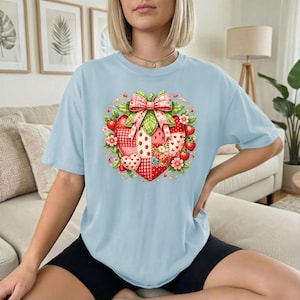 Può includere: Una t-shirt azzurro chiaro con una grande grafica centrale a forma di cuore composta da fragole. Il design presenta un motivo patchwork a quadretti rossi e bianchi, pois e piccole fragole, sormontato da un grande fiocco rosa e bianco.