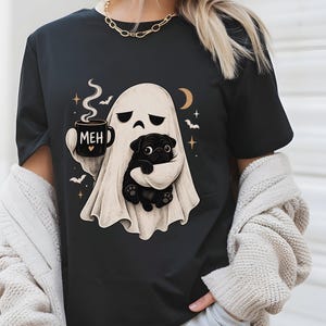 May include: A black t-shirt featuring a graphic of a white ghost holding a black mug with the text MEH and a small heart. The ghost is embracing a small black pug dog, surrounded by small bats, stars, and a crescent moon in a beige color palette.