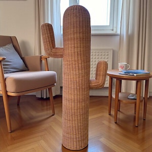 May include: A tall, woven rattan cactus-shaped coat rack. The cactus has a cylindrical base and two arm-like extensions. The rack is a light brown color and stands on a wooden floor. A chair and small table are also in the room.