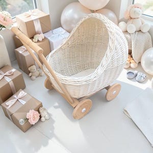 Könnte beinhalten: Ein weißer Puppenwagen aus Korbgeflecht mit Holzrädern und einem Griff. Der Wagen ist umgeben von Geschenkboxen mit rosa Bändern, einem Stofftier und Luftballons. Die Szene spielt auf einer weißen Oberfläche.
