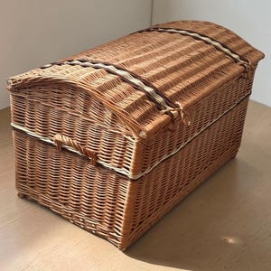 May include: A woven wicker chest with a domed lid. The chest is a warm brown color with a lighter-colored trim and a clasp closure. The chest is sitting on a wooden surface.