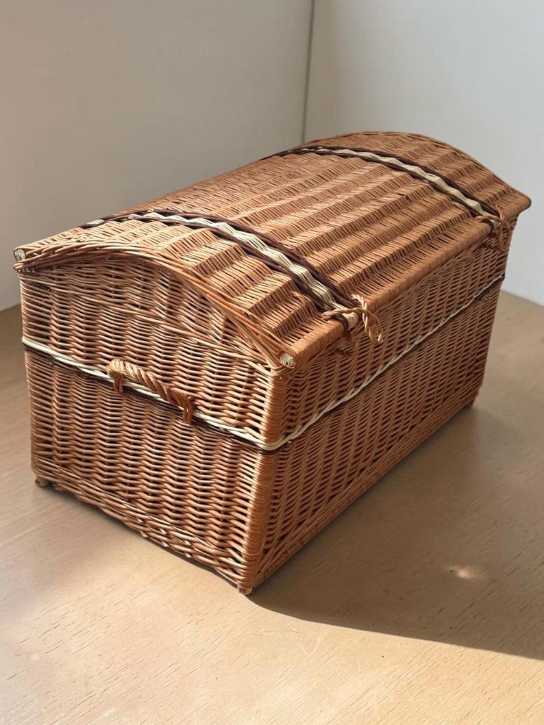 May include: A woven wicker chest with a domed lid. The chest is a warm brown color with a lighter-colored trim and a clasp closure. The chest is sitting on a wooden surface.
