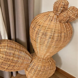 May include: A large, woven wicker cactus sculpture in natural tones. The sculpture features a rounded base, a central body, and three smaller, rounded protrusions at the top, resembling cactus pads. The woven texture is visible throughout the design.