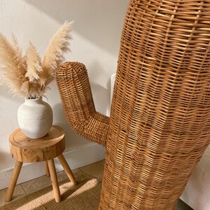 May include: A large, tall, brown woven wicker cactus sculpture with a single side arm. Next to it sits a small, round, light-colored wooden stool with three legs, holding a white ceramic vase filled with fluffy, beige pampas grass plumes.