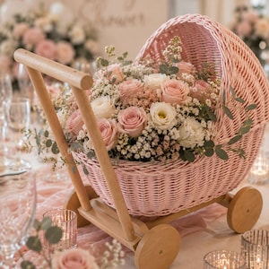 May include: A light pink woven wicker baby carriage centerpiece filled with a floral arrangement of light pink roses, white roses, baby's breath, and green eucalyptus leaves, featuring a light wooden handle and four small wooden wheels.