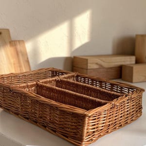 May include: A rectangular, woven wicker cutlery organizer with multiple compartments. The natural brown basket is designed to hold silverware and utensils. The basket is sitting on a white surface.