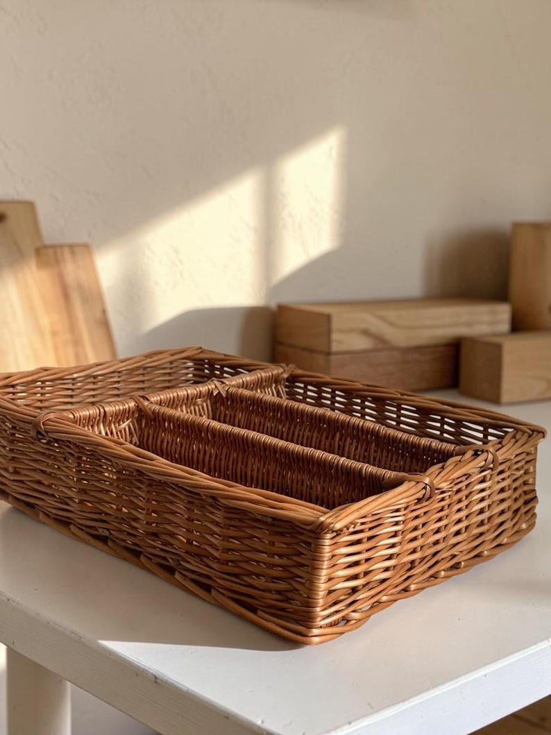 May include: A rectangular, woven wicker cutlery organizer with multiple compartments. The natural brown basket is designed to hold silverware and utensils. The basket is sitting on a white surface.