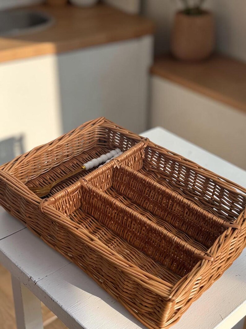 May include: A rectangular woven wicker cutlery tray with four compartments. The tray is a warm brown color and contains a gold-colored fork with a white handle. The tray is designed to organize silverware and utensils.