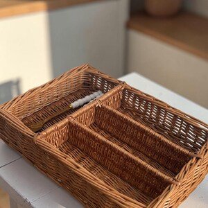 May include: A rectangular, woven wicker cutlery organizer with four compartments. The natural brown basket holds a gold-colored fork with a white handle. The organizer sits on a white surface, with a blurred kitchen background.