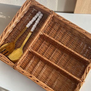 May include: A woven wicker cutlery tray with multiple compartments. Two gold-colored serving utensils rest inside the tray. The tray is brown and sits on a white surface. The serving utensils have white beaded handles.