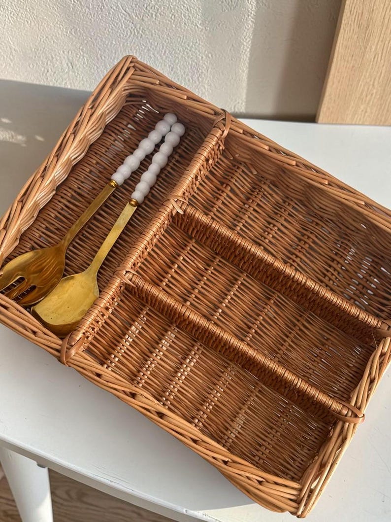 May include: A woven wicker cutlery tray with multiple compartments. Two gold-colored serving utensils rest inside the tray. The tray is brown and sits on a white surface. The serving utensils have white beaded handles.