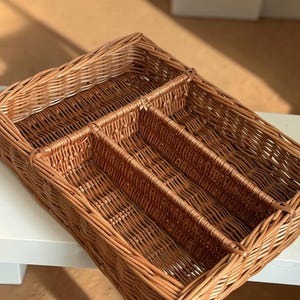 May include: A rectangular, woven wicker basket with multiple compartments. The basket is a warm brown color and appears to be handmade. It is designed for organizing items and has a natural, rustic aesthetic.
