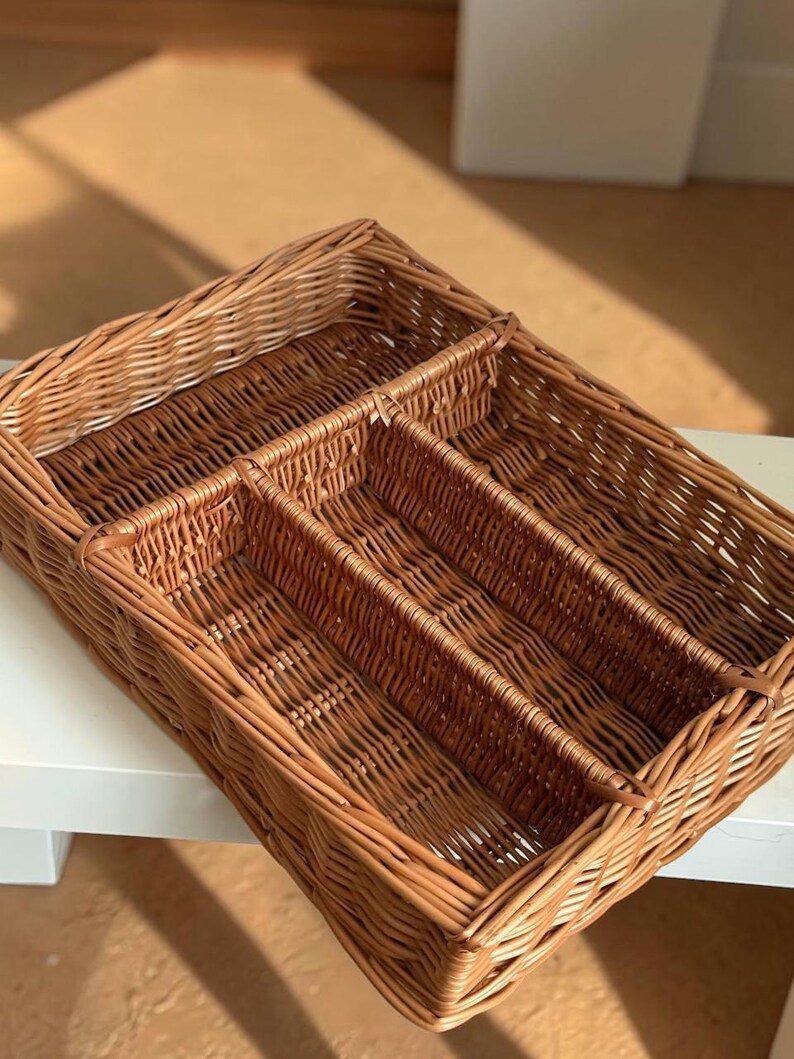 May include: A rectangular, woven wicker basket with multiple compartments. The basket is a warm brown color and appears to be handmade. It is designed for organizing items and has a natural, rustic aesthetic.