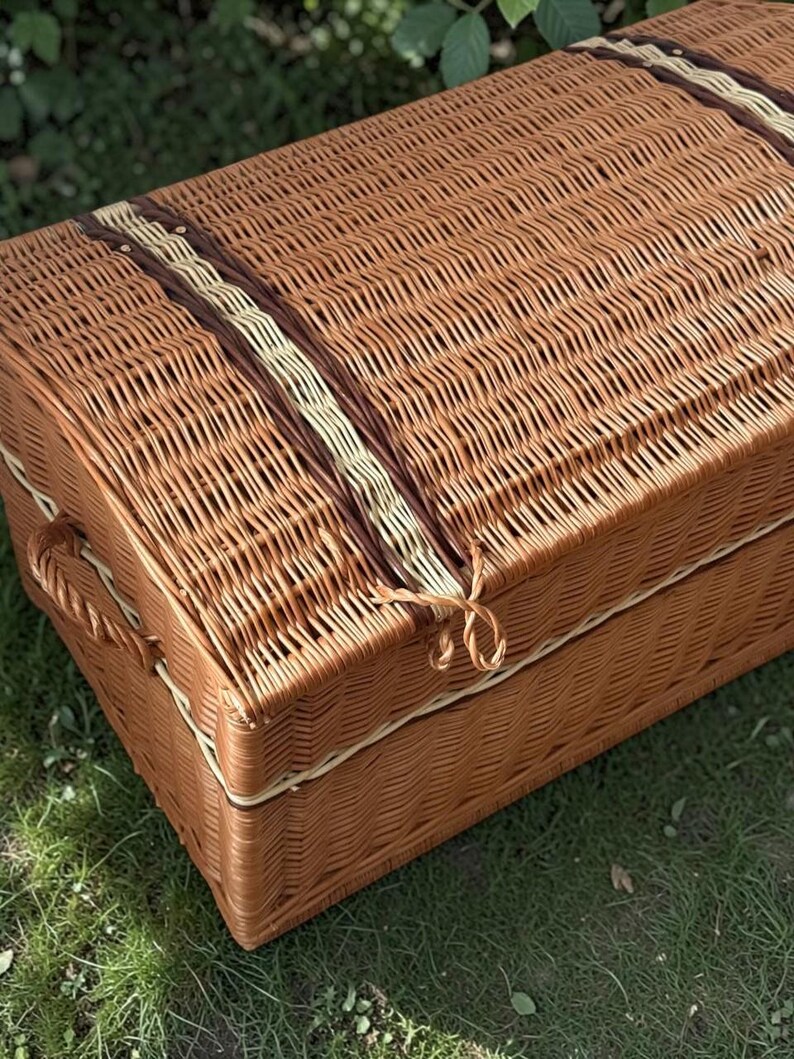 May include: A woven wicker trunk in shades of brown, with a curved top and a rectangular shape. The trunk features decorative bands of darker brown and cream-colored wicker. The trunk is sitting on green grass.