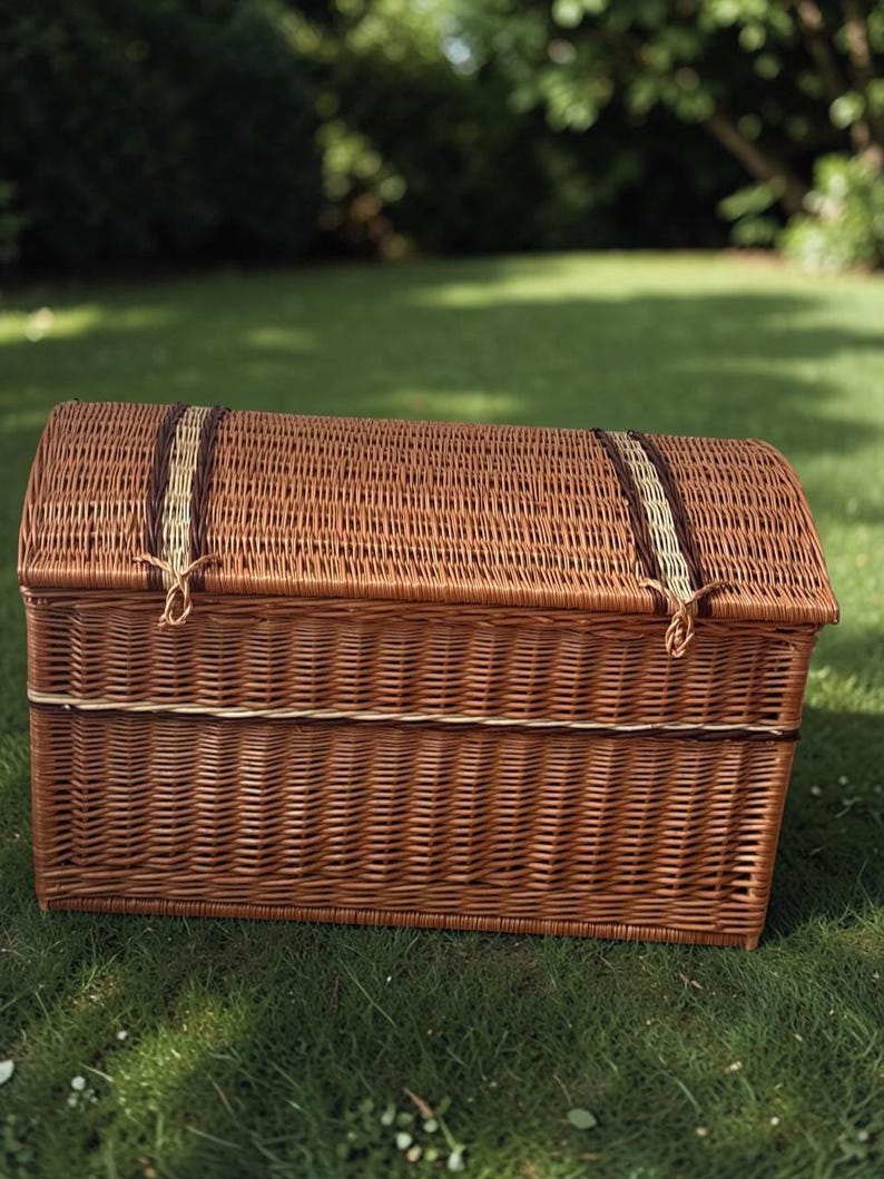 May include: A brown wicker trunk with a domed lid, resting on green grass. The trunk has a rectangular base and is secured with two straps across the top. The straps are a mix of brown and tan. The trunk is a classic storage solution.