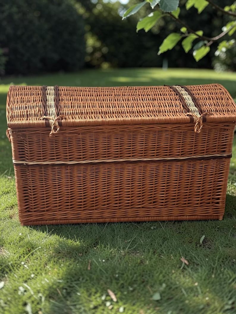 May include: A brown wicker chest with a curved top and a rectangular base. The chest has a woven design with a lighter-colored band around the middle and on the lid. The chest is sitting on green grass.