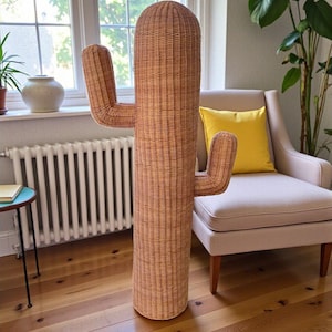 May include: A tall, woven rattan cactus sculpture with two side arms. The cactus is a light brown color and stands on a wooden floor. The sculpture is placed near a window and a beige armchair with a yellow pillow.
