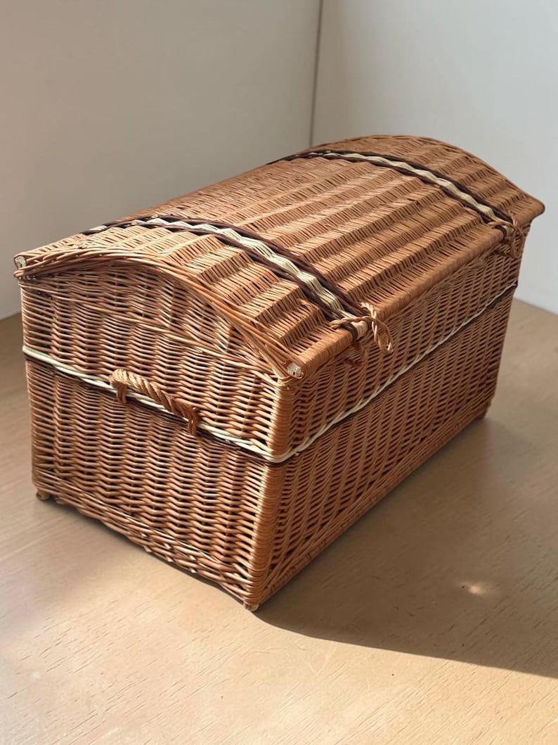 May include: A woven wicker chest with a domed lid. The chest is a warm brown color with a lighter-colored trim and a clasp closure. The chest is sitting on a wooden surface.