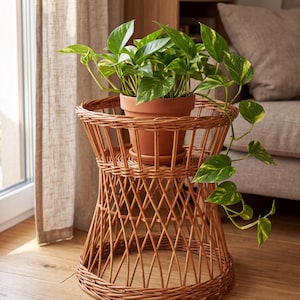 Op de afbeelding: Een lichtbruine gevlochten rieten plantenstandaard met een uitlopende zandlopervorm en een rond blad. Op de standaard staat een terracotta-kleurige pot met een groene pothosplant met geel gevlekte bladeren die langs de zijkant hangen.