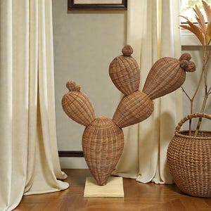 May include: A large, brown wicker cactus sculpture with multiple rounded sections, standing on a small wooden base. The sculpture is placed on a wooden floor, with beige curtains and a framed picture in the background. A wicker basket with a plant is also visible.