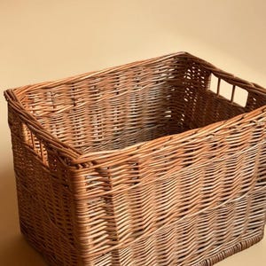 May include: A rectangular woven wicker basket in shades of brown and tan. The basket has a sturdy construction with a woven pattern and cut-out handles on the sides. The basket is empty and sits against a neutral background.