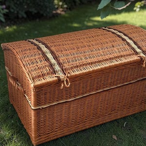 May include: A large, woven wicker trunk in shades of brown. The trunk has a rounded top and is secured with woven straps and rope closures. The trunk is sitting on green grass, suggesting outdoor use. The trunk is likely used for storage.