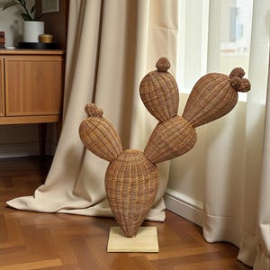 May include: A large, decorative cactus sculpture made of woven wicker. The sculpture is brown and stands on a small, square wooden base. The cactus has multiple pads and a rounded top, resembling a prickly pear cactus.