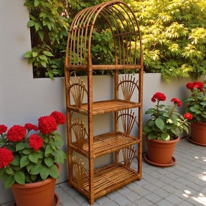 May include: A brown wicker plant stand with an arched top and three shelves. The stand is decorated with fan-shaped accents. Red flowering plants in terracotta pots are placed around the stand, creating a garden setting.