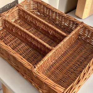 May include: Woven wicker cutlery tray with three compartments. The rectangular tray is a warm brown color and is designed to organize silverware. The tray is made of natural materials and is suitable for kitchen organization.