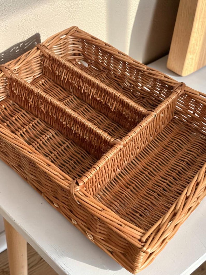 May include: Woven wicker cutlery tray with three compartments. The rectangular tray is a warm brown color and is designed to organize silverware. The tray is made of natural materials and is suitable for kitchen organization.