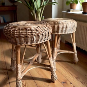 May include: Two round, woven wicker stools with light brown wooden legs. The stools have a natural, neutral color palette and are placed on a wooden floor. The stools are suitable for indoor use and add a touch of rustic charm to any space.